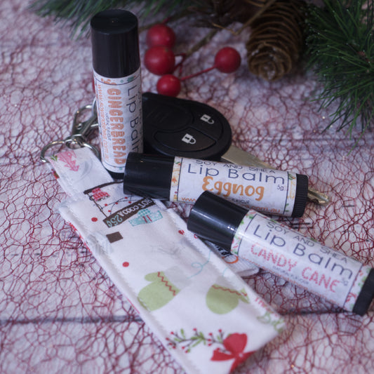 Soy and Shea's Merry & Bright CLip Balm + Lip Balm Bundle featuring limited edition holiday-themed scents like gingerbread, eggnog, and candy cane is beautifully showcased against a festive backdrop adorned with pine branches and berries. The display is complete with a camera lens cap and a fabric pouch embellished with holiday prints, enhancing the scene's charm.