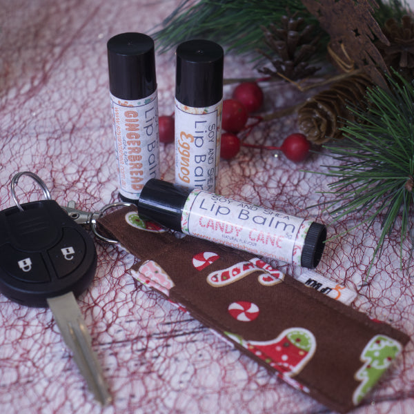 The Sugar & Spice CLip Balm + Lip Balm Bundle by Soy and Shea features three festive lip balms named "Gingerbread," "Eggnog," and "Candy Cane." They sit elegantly on a surface decorated with pine needles and red berries, accompanied by a car key and a holiday-themed case that enhance the seasonal ambiance.