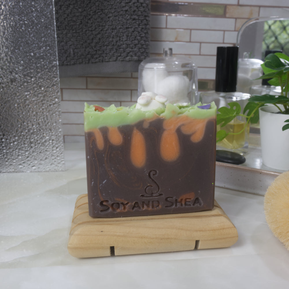 A soap bar stands on a wooden rack on a bathroom bench along with perfume, jars and a plant.   The soap features a brown base layer with bronze swirls and orange drops made to look like carrots.  The top has a layer of bright green and is spiked to look like grass.  The top has a soap decoration of a bunny butt with little pink paw pads.