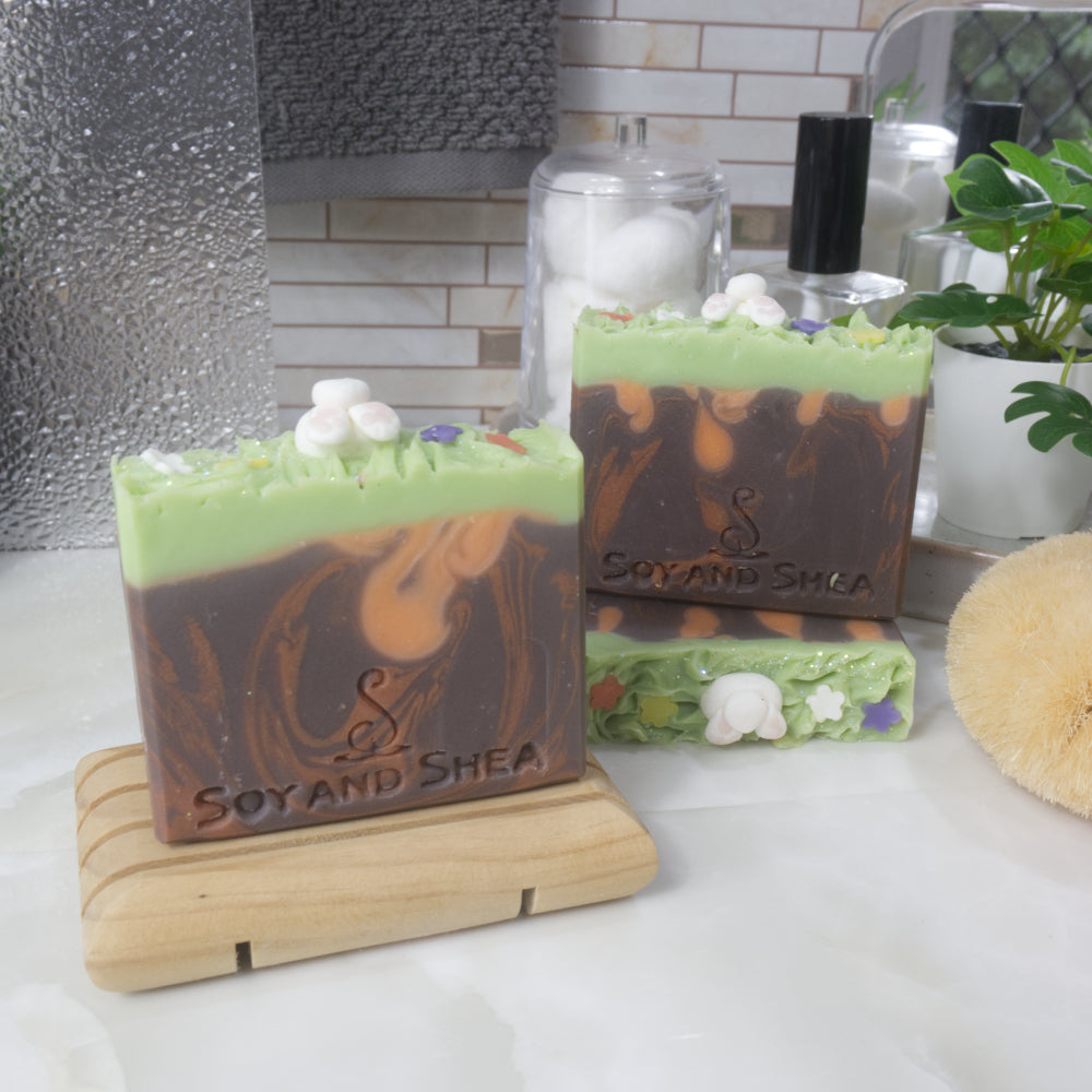 A selection of three soaps displayed on a bathroom bendch along with a bottle of perfume, plant and jar of cotton balls.  One soap sits on a wooden rack to the front left of the image, another two are displayed behind with one laying flat showing the top of the soap with a third standing on top. The soaps are brown with bronze swirls and orange drops.  A layer of green soap finishes the top and looks like grass.  Each soap had a little bunny butt decoration along with small flowers