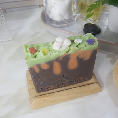 A soap bar stands on a wooden rack on a bathroom bench along with perfume, jars and a plant.   The soap features a brown base layer with bronze swirls and orange drops made to look like carrots.  The top has a layer of bright green and is spiked to look like grass.  The image focuses on the top which has a soap decoration of a bunny butt with little pink paw pads made of soap along with some soap flowers