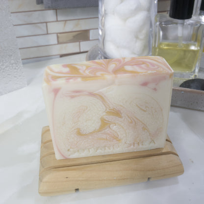 A bar of soap sits on a wooden soap dish on a bathroom countertop surrounded by perfume and a jar of cotton balls.   The soap has a white base colour with swirls of pink and gold throughout and across the top.