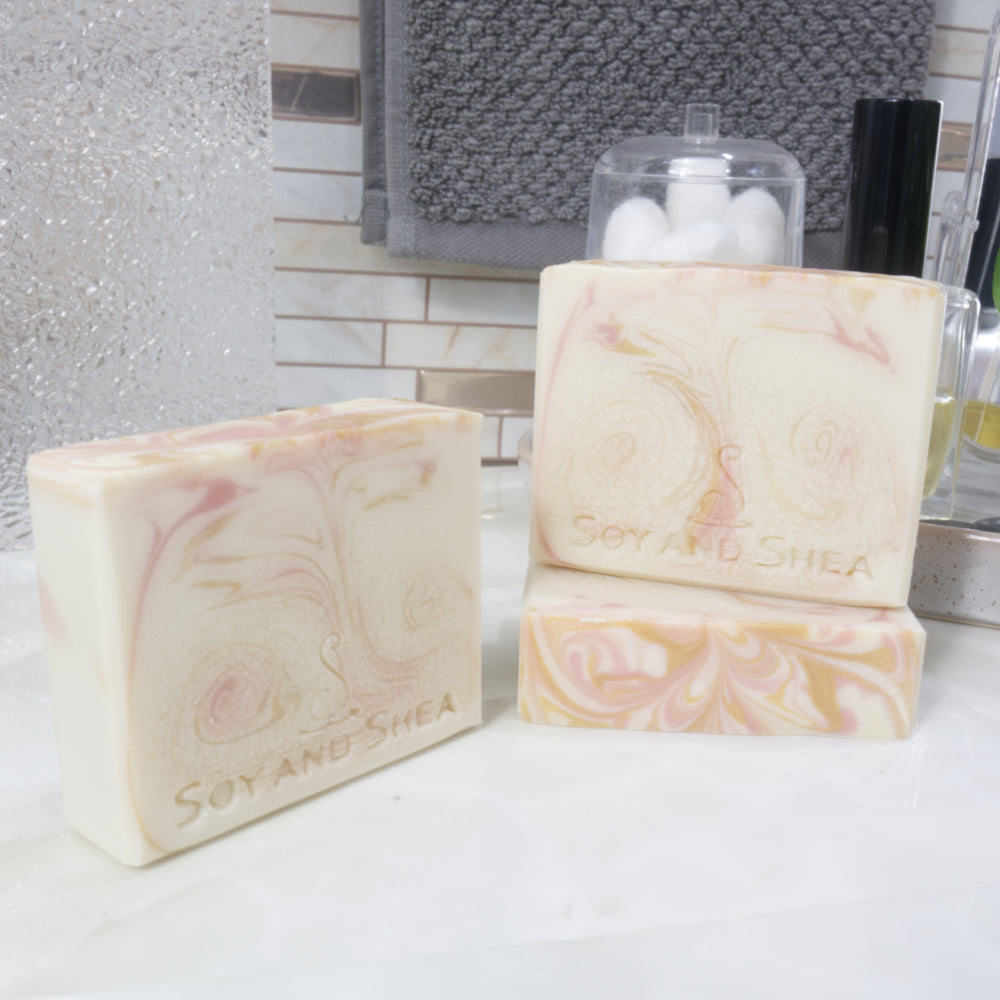 Three rectangular soaps are presented on a bathroom counterstop. To the left, a soap bar stands upright on the bench, while to the right, another lays flat showing the flat top with decorative colour swirl while another stands upright on it.  All the soaps are a cream base colour with varying swirls of pink and gold 