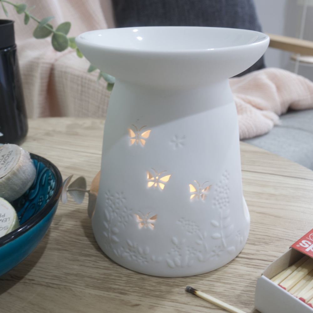 A close image of a matte white porcelain fragrance warmer sitting on a light wooden table next to a blue bowl of wax melts and a box of matches. The warmer features embossed botanical details at the base and lit butterfly cutouts that glow warmly from an internal tealight candle. In the blurred background, a pink textured throw blanket and a dark grey cushion rest on a chair