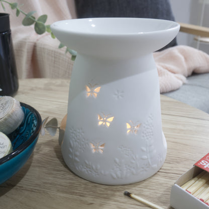 A close image of a matte white porcelain fragrance warmer sitting on a light wooden table next to a blue bowl of wax melts and a box of matches. The warmer features embossed botanical details at the base and lit butterfly cutouts that glow warmly from an internal tealight candle. In the blurred background, a pink textured throw blanket and a dark grey cushion rest on a chair