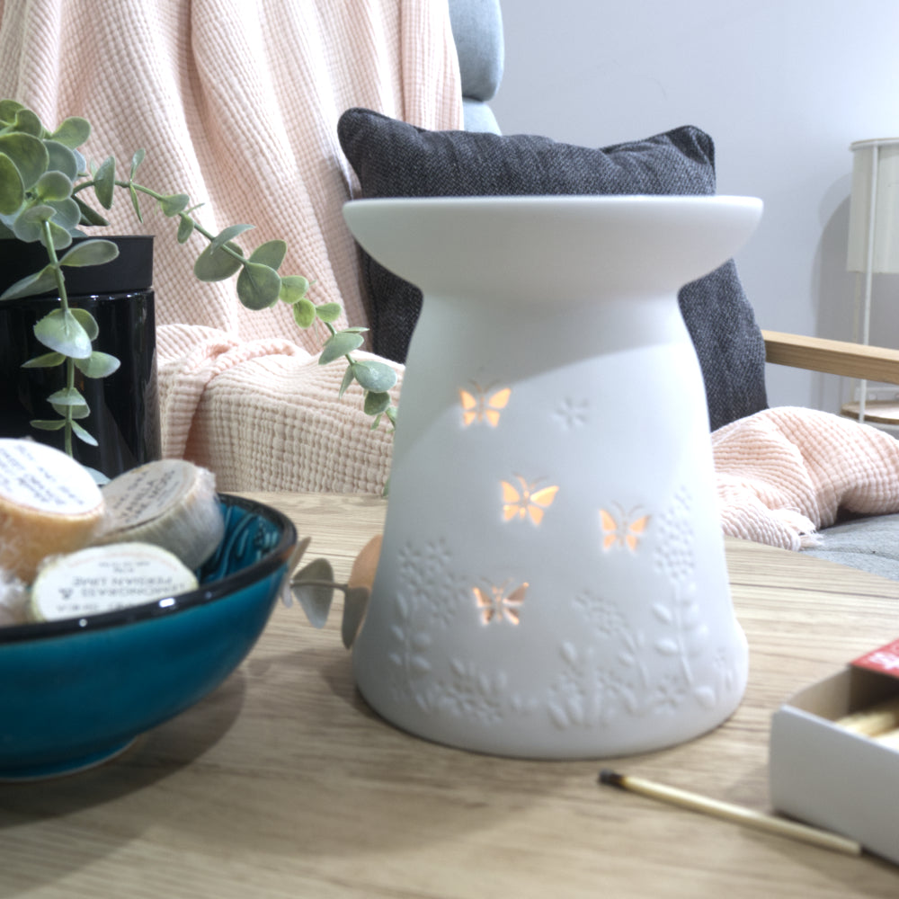 A matte white porcelain fragrance warmer sits on a light wooden table next to a blue bowl of wax melts and a box of matches. The warmer features embossed botanical details at the base and lit butterfly cutouts that glow warmly from an internal tealight candle. In the blurred background, a pink textured throw blanket and a dark grey cushion rest on a chair