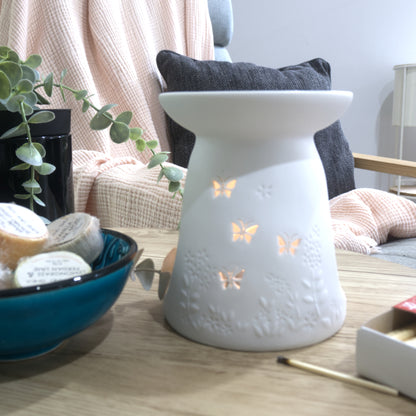 A matte white porcelain fragrance warmer sits on a light wooden table next to a blue bowl of wax melts and a box of matches. The warmer features embossed botanical details at the base and lit butterfly cutouts that glow warmly from an internal tealight candle. In the blurred background, a pink textured throw blanket and a dark grey cushion rest on a chair