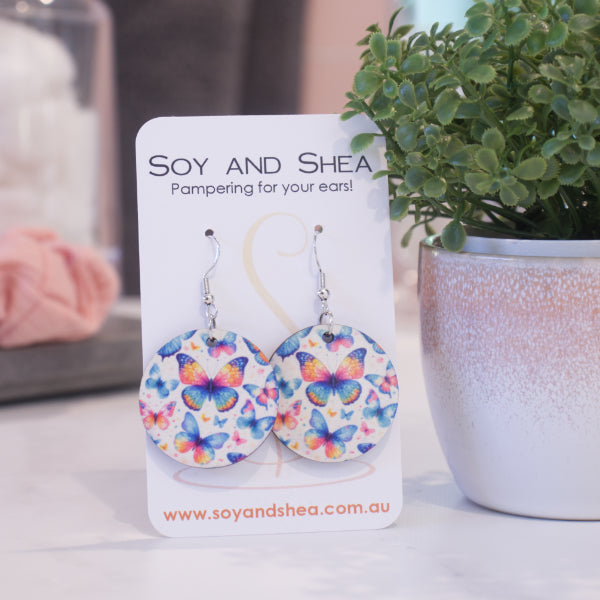 A pair of earrings on silver hooks hang on a white card branded with SOY AND SHEA leaning against a pot with a green leafy plant and a blurred background.  The earrings are round with a random scattering of butterflies in shades of blue, pink and orange.