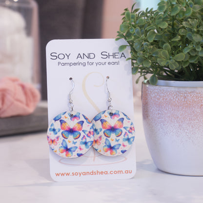 A pair of earrings on silver hooks hang on a white card branded with SOY AND SHEA leaning against a pot with a green leafy plant and a blurred background.  The earrings are round with a random scattering of butterflies in shades of blue, pink and orange.