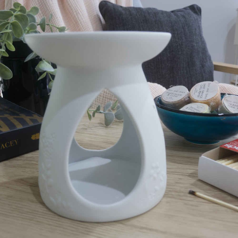 A matte white porcelain fragrance warmer sits on a light wooden table next to a blue bowl of wax melts and a box of matches. The image shows the warmer from the side showing large teardrop openings on the side of the warmer.