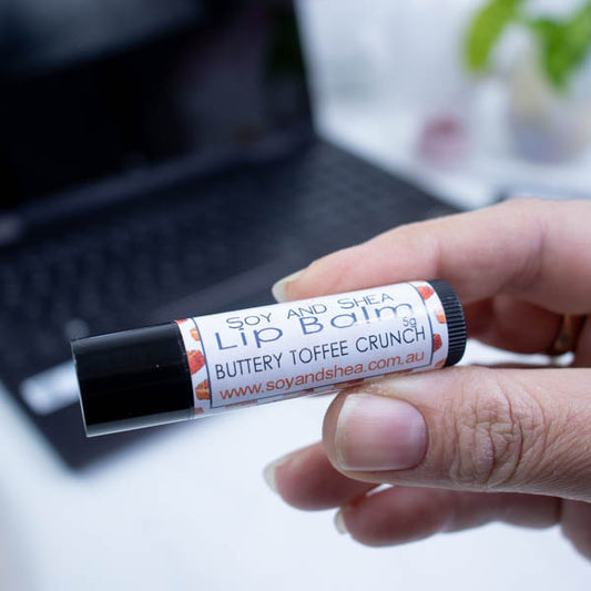 A hand holds a black tube of lip balm against a blurred background with an open laptop computer on a white surface.  The label has text LIP BALM BUTTERY TOFFEE CRUNCH on a white background with a border showing square toffees.
