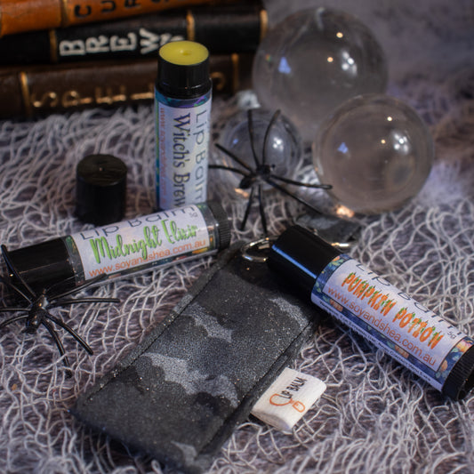 An arrangement of Soy and Shea’s Bats Clip Balm + Lip Balm Bundle, labeled "Witch's Brew," "Midnight Kiss," and "Pumpkin Potion," rests on a surface draped with white web-like material. Nearby are small black plastic spiders, clear spherical ornaments, and a few tiny accessories. The scene is completed with books in the background.