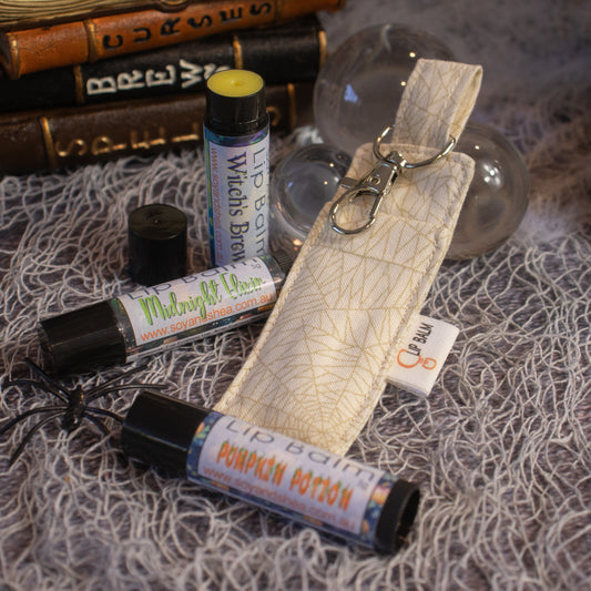 Three tubes of Soy and Shea lip balm labeled "Witch's Brew," "Midnight Moon," and "Pumpkin Potion" rest on a textured gray surface. Beside them is a Cobwebs Clip Balm holder with a keychain clip. In the background, there are stacked books, clear round ornaments, and a woven gray cloth.