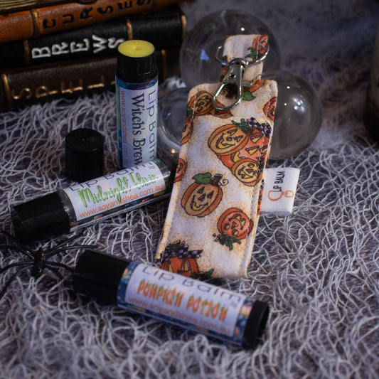 The "Pumpkin Patch Clip Balm + Lip Balm Bundle" from Soy and Shea features Halloween-themed lip balms, such as "Witch's Brew" and "Pumpkin Potion," displayed with a Clip Balm holder decorated with pumpkin faces. This arrangement is set against textured fabric and books, creating an atmosphere reminiscent of a spooky Pumpkin Patch.