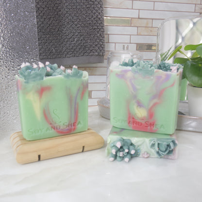 Three soap bars are displayed on a bathroom countertop with a towel and plant.  One soap lays flat on the bench showing a selection of green cacti decorating the top.  Another stands upright on it, with a third standing on a wooden soap dish to the left.  All soap have a bright green base colour, with varying swirls in red, yellow and purple.  Each soap has a different selection of cacti embellishments.
