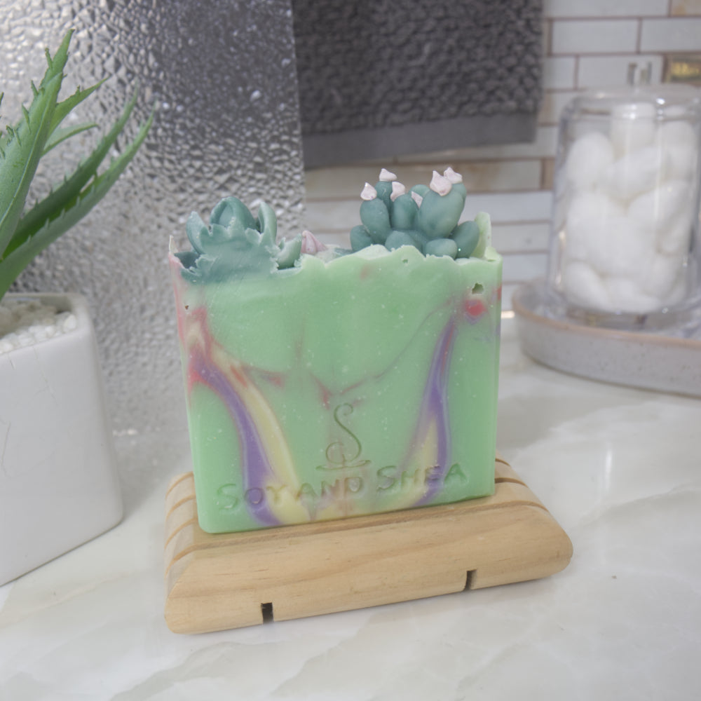 A soap bar standing on a wooden soap rack with a blurred background.  The soap is bright green with swirls of purple, yellow and red.  On the top of the soap is a selection of Cacti embellishments.