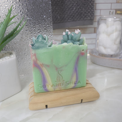 A soap bar standing on a wooden soap rack with a blurred background.  The soap is bright green with swirls of purple, yellow and red.  On the top of the soap is a selection of Cacti embellishments.