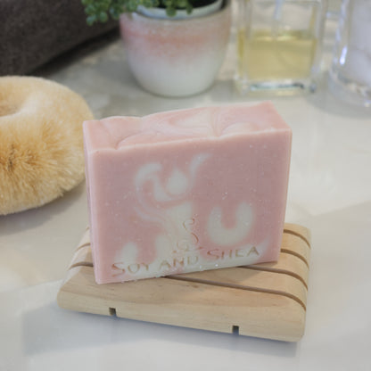 An overhead shot showing a pale pink soap bar with white swirls sitting on a wooden soap dish on a bathroom counter top.  In the background a body brush and bottle of perfume can be seen.