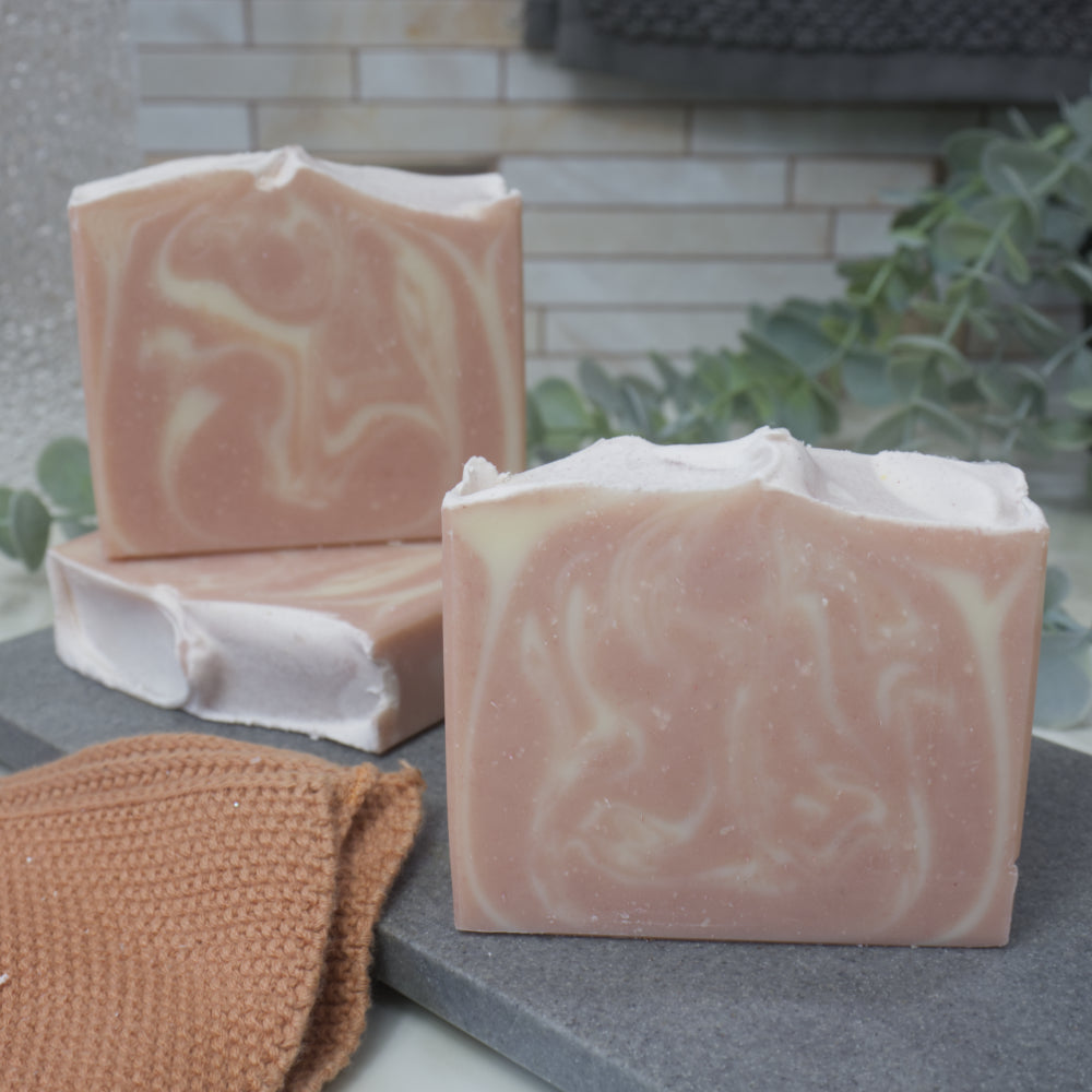 Three bars of soap with a marbled pattern of pink and white sit on a stone surface, with a brown face wash and greenery in the background.