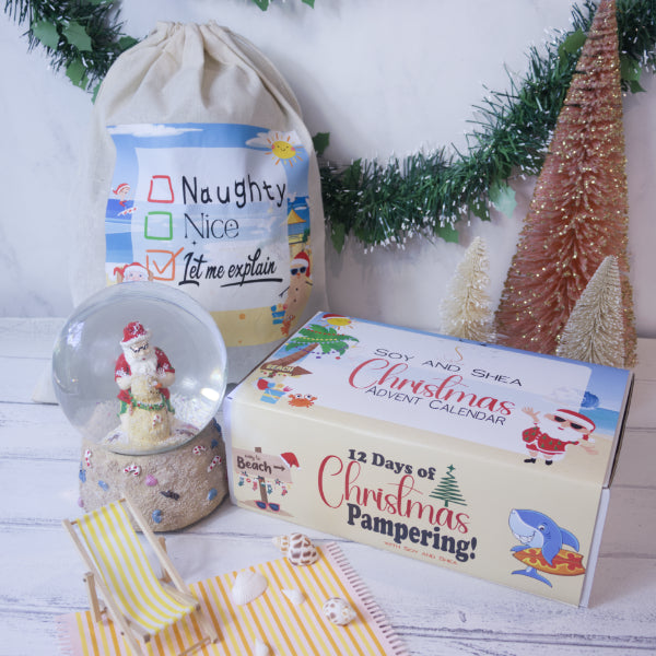 For an Aussie Summer Christmas celebration, feature a "Naughty or Nice" drawstring bag, the "12 Days of Christmas Pampering" advent calendar from Soy and Shea—available in a vegan option—and a Santa snow globe lounging on a mini beach chair. The holiday decorations are completed with green garland and small gold trees.