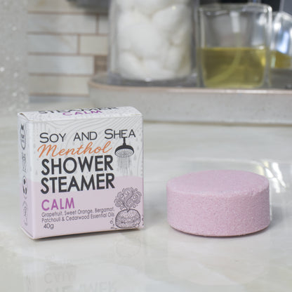 A boxed shower steamer and the steamer itself on a countertop in a bathroom setting. The box reads 'Soy and Shea Menthol Shower Steamer Calm' and the round steamer is a pale mauve color.