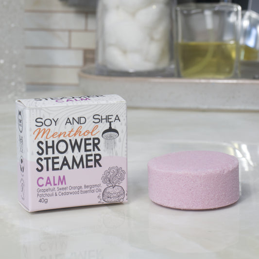 A boxed shower steamer and the steamer itself on a countertop in a bathroom setting. The box reads 'Soy and Shea Menthol Shower Steamer Calm' and the round steamer is a pale mauve color.