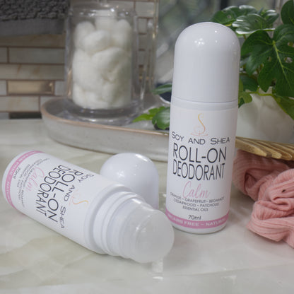 Two bottles of Roll-On Deodorant are displayed on a bathroom counter top with a jar of cotton balls, hair tie and greenery.  One bottle stands upright with a domed cap, while another lays flay with the cap removed showing a ball roller in the top