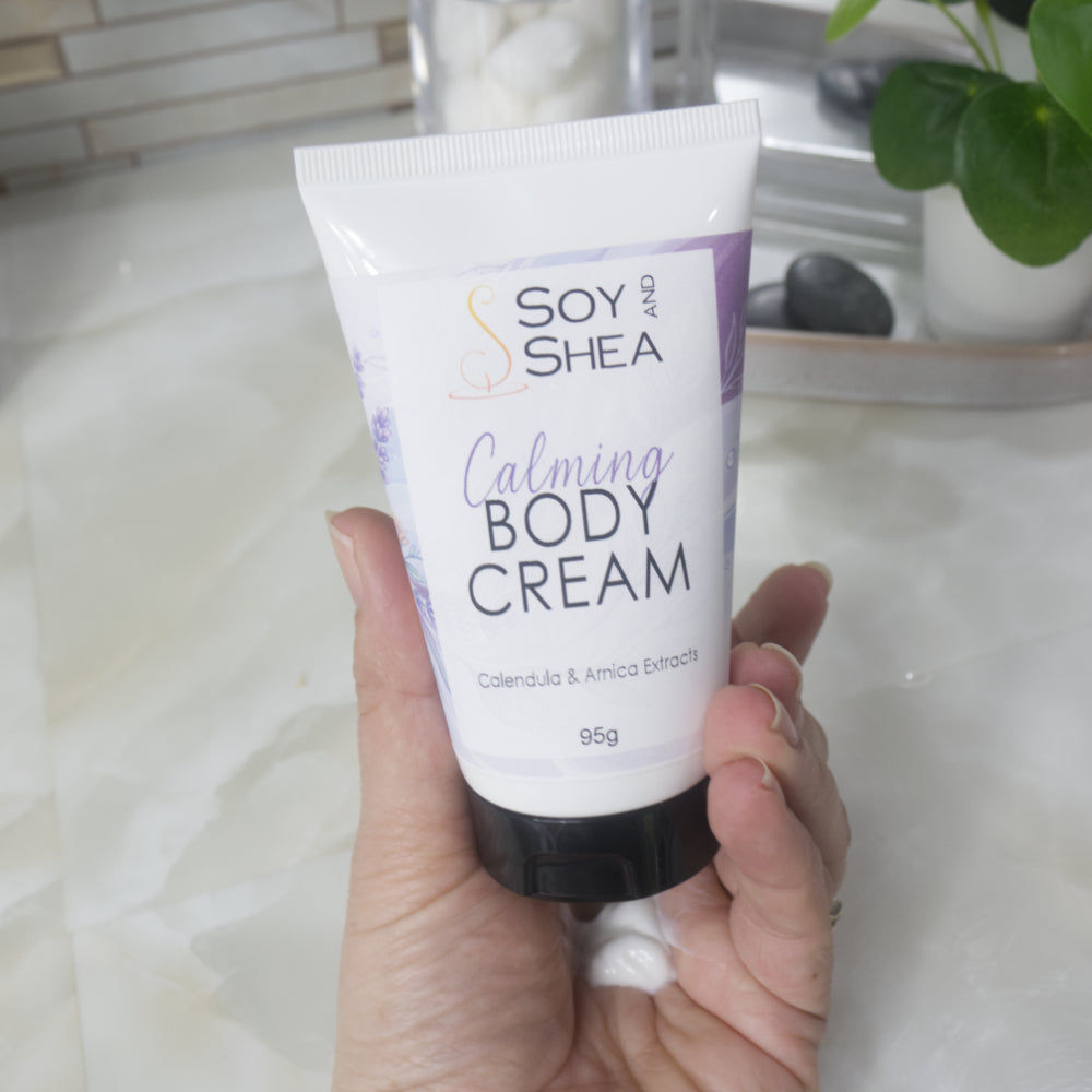 A hand holds a white tube with black cap labelled as Calming Body Cream with calendula and Arnica Eztracts.  In the palm of the hand is a dollop of white cream that has been squeezed out of tube. 