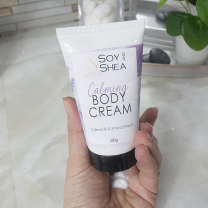 A hand holds a white tube with black cap labelled as Calming Body Cream with calendula and Arnica Eztracts.  In the palm of the hand is a dollop of white cream that has been squeezed out of tube. 