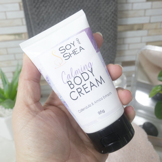 A hand holding a squeeze tube of Soy of Shea Calming Body Cream with a white body, purple text and accents, and a black cap, set against a softly blurred bathroom background.