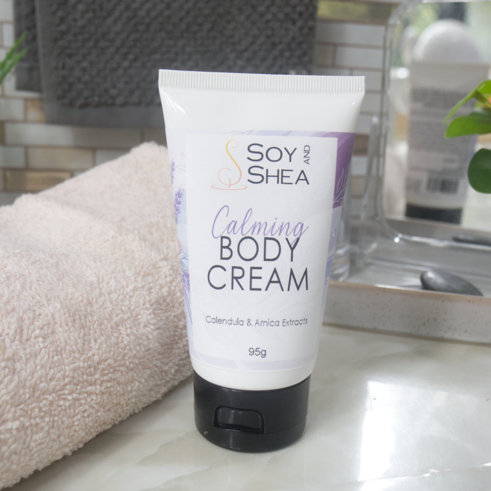 A white tube with black flip top cap stands on a bathroom counter next to a white towel and bathroom accessories.  The tube features a decorative product label with blue background announcing it as Calming Body Cream with calendula, and Arnica Extracts