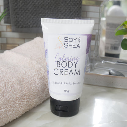 A white tube with black flip top cap stands on a bathroom counter next to a white towel and bathroom accessories.  The tube features a decorative product label with blue background announcing it as Calming Body Cream with calendula, and Arnica Extracts