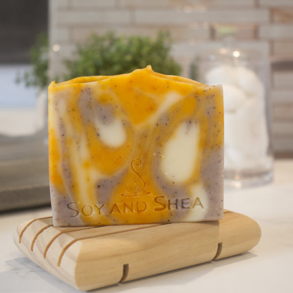 A bar of soap with a swirl pattern featuring orange, white, and purple colors, resting on a wooden soap dish against a blurred background. The soap has 'SOY AND SHEA' branded into its top surface.