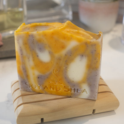 A bar of soap with a swirl pattern featuring orange, white, and purple colors, resting on a wooden soap dish against a blurred background. The soap has 'SOY AND SHEA' branded into its top surface.