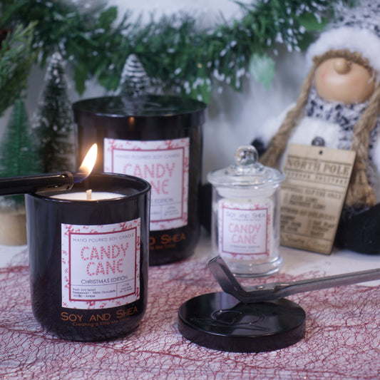 A cozy scene featuring the "Candy Cane Soy Candle (Christmas Edition)" from Soy and Shea, glowing warmly on a textured surface. Infused with delightful fragrance oils, the candles fill the air, while holiday decorations like a miniature tree and snowman spread festive cheer. A candle lid and wick trimmer are prominently displayed in the foreground.