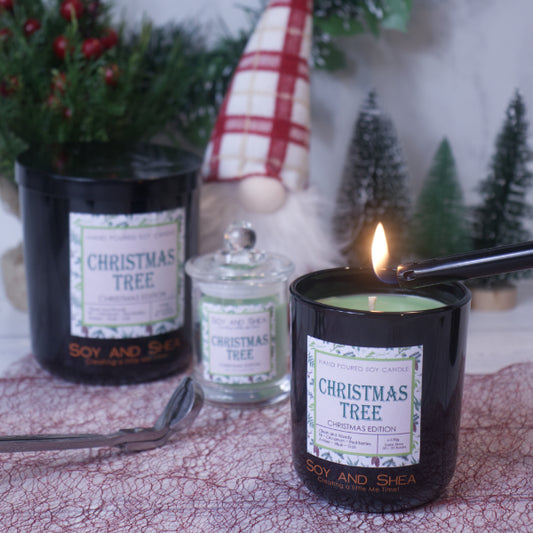 A cozy holiday scene featuring a lit "Christmas Tree Soy Candle (Christmas Edition)" by Soy and Shea, infused with premium fragrance oils in a sleek black jar, surrounded by closed candles and festive decorations. A small evergreen tree and a Santa figure can be seen in the background, all set on a lace-textured surface.