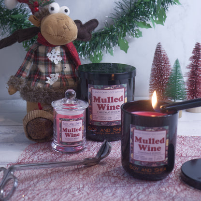 A festive scene is set with Soy and Shea's Christmas Edition Mulled Wine Soy Candle, accompanied by two others from the same collection. Each candle features natural cotton wicks, releasing a warm mulled wine fragrance into the air. In the background, a plush reindeer toy, decorative trees, and a pine garland enhance the winter-themed table setting.