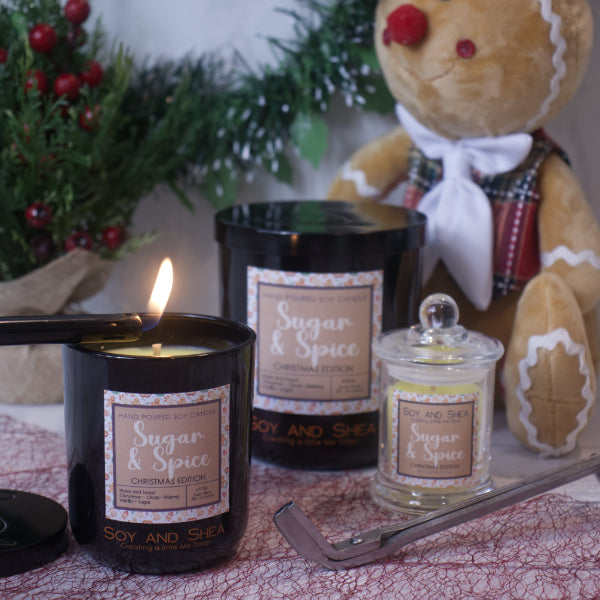 A cozy scene showcases a selection of both lit and unlit Sugar & Spice Soy Candle (Christmas Edition) by Soy and Shea, accompanied by a gingerbread-themed teddy bear. A wick trimmer sits close by, surrounded by a festive backdrop featuring pine branches and berries, ensuring a non-toxic burn with a warm holiday fragrance.