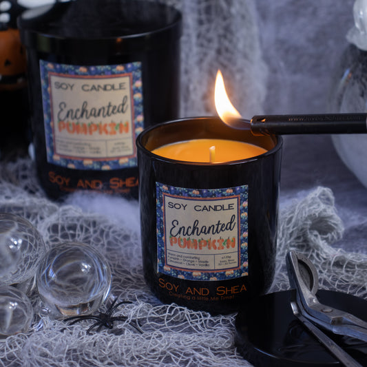 A lit Enchanted Pumpkin Soy Candle (Halloween Edition) from Soy and Shea flickers to life with a match. Another unlit soy candle, crafted with premium fragrance oils, sits in the background among cobwebs and glass orbs, enhancing a spooky autumn vibe. Nearby, eco-friendly wick trimmers rest gracefully.