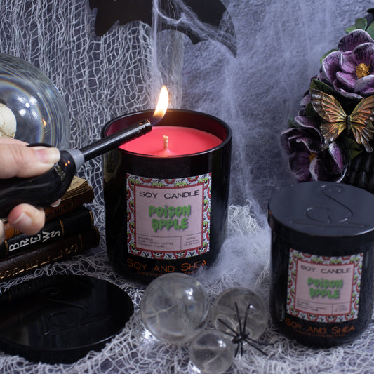 A hand lights a "Poison Apple Soy Candle (Halloween Edition)" by Soy and Shea with a lighter. This eco-friendly candle is housed in a black jar adorned with a pink label. Decorative items such as purple flowers, clear glass orbs, and a spider figure surround it, all resting on a surface covered with spider webs.