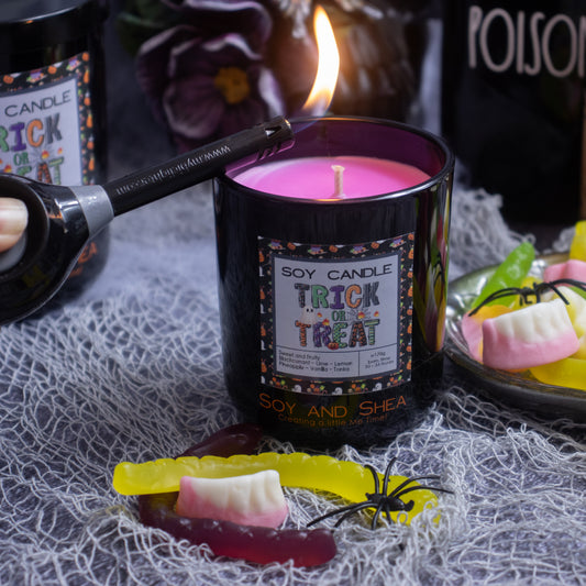 A hand is lighting a Trick or Treat Soy Candle (Halloween Edition) by Soy and Shea, featuring blackcurrant-scented bright pink wax. It's positioned on a gray netting cloth surrounded by gummy candies shaped like fangs and worms, creating an eco-friendly Halloween atmosphere with spooky props in the background.