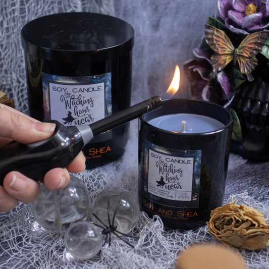 A hand is lighting an eco-friendly black candle labeled "Witching Hour Soy Candle (Halloween Edition)" by Soy and Shea with a lighter, next to another unlit candle in a similar jar. The scene includes Halloween-themed decorations like a black ceramic skull, dried flowers, a fake spider, and a web-covered surface.