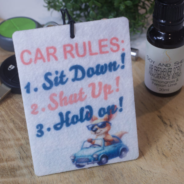A car air freshener stands upright on a wood surface with a bottle of scent and car key.  The Air freshener has text that reads CAR RULES.  SIT DOWN.  SHUT UP. HOLD ON.  In the lower right corner is a cartoon image of a large kangaroo in a small blue car without a roof.