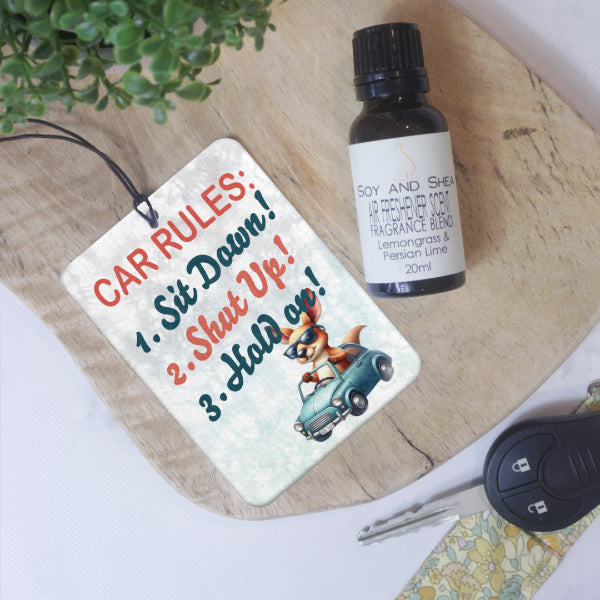 A rectangular car air freshener lies on a wooden surface next to a glass bottle of Air Freshener Scent and car key.  The Air Freshener has a blue mottle background with the Text CAR RULES across the top in red.  Below are three rules including  SIT DOWN SHUT UP HOLD ON.  In the lower right corner is an image of a large kangaroo driving a small blue car without a roof