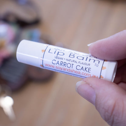 A hand holds a white tube labelled lip balm over a blurred image of an open handbag with contents spilling onto the table.  The lip balms features a white background with small illustrations of cake slices