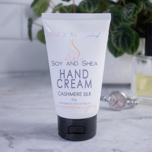 A tube of Cashmere Silk Hand Cream from Soy and Shea is placed on a marble surface with a tiled background.  The tube features a label with white background adorned with subtle leaf illustrations, accompanied by text reading HAND CREAM. In the blurred background, a wristwatch, perfume bottle, and houseplant can be seen.