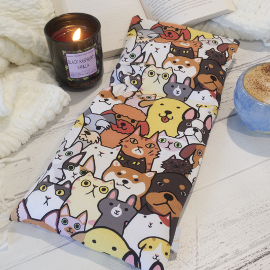 A long retangular heat pack lying on a flat surface and touching an open book, with a burning candle and a cup in the background.  The heat pack features a design of various breeds of cats and dogs sitting side-by-side and in rows.  It is a bold pattern where the animals are outlined in black and coloured in bold earthy colours