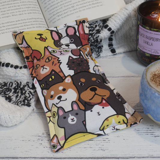 A Cats and Dogs Mini Heat Pack rests against a black and white blanket with open book, next to a burning candle and coffee cup creating a cosy scene.   The Heat Pack feature a drawing of various cats and dogs sitting side by side and in rows.  Each animal has a black outlined filled with bold natural colours