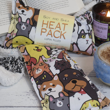 Two Cats and Dogs Mini Heat Pack by Soy and Shea rests near a ceramic coffee cup, a burning candle, and an open book—creating an inviting cosy scene.  The Mini Heat packs feature a variety of hand-drawn cats and dog sitting side by side and in rows.  They are bright bold colours and all outlined in black.  One pack shows the design horizontally whereas the other is vertical.