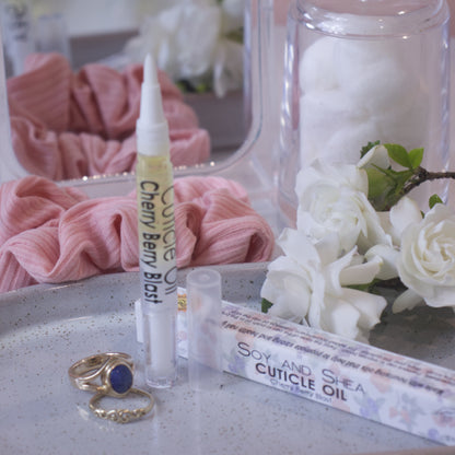 A clear, narrow tube with the lid removed showing a brush tip stands upright on a stone tray.  Beside it sits a cardboard box labelled CUTICLE OIL and a set of gold rings.  In the background is a bathroom counter top with a pink scrunchie, small mirror, jar of cotton balls and a some white  flowers.